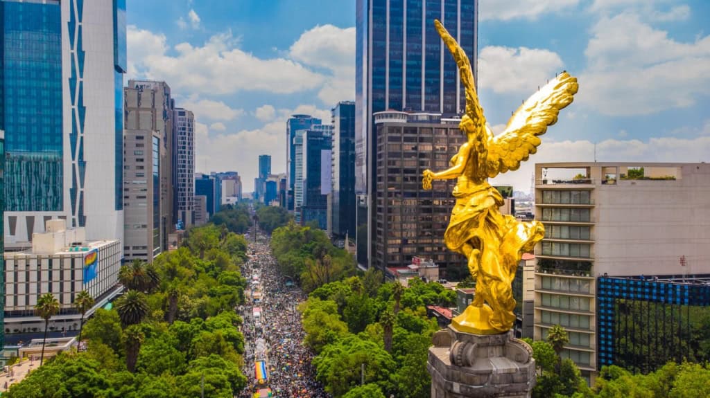 An aerial view of Mexico City showcasing the Angel of Independence monument amidst modern skyscrapers and lush green parks on a bright, sunny day.