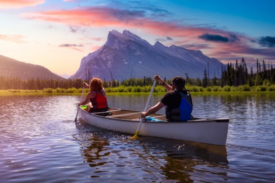 Two people paddling a canoe on a serene lake with mountain views at sunset, representing outdoor activities and natural beauty in Canada.
