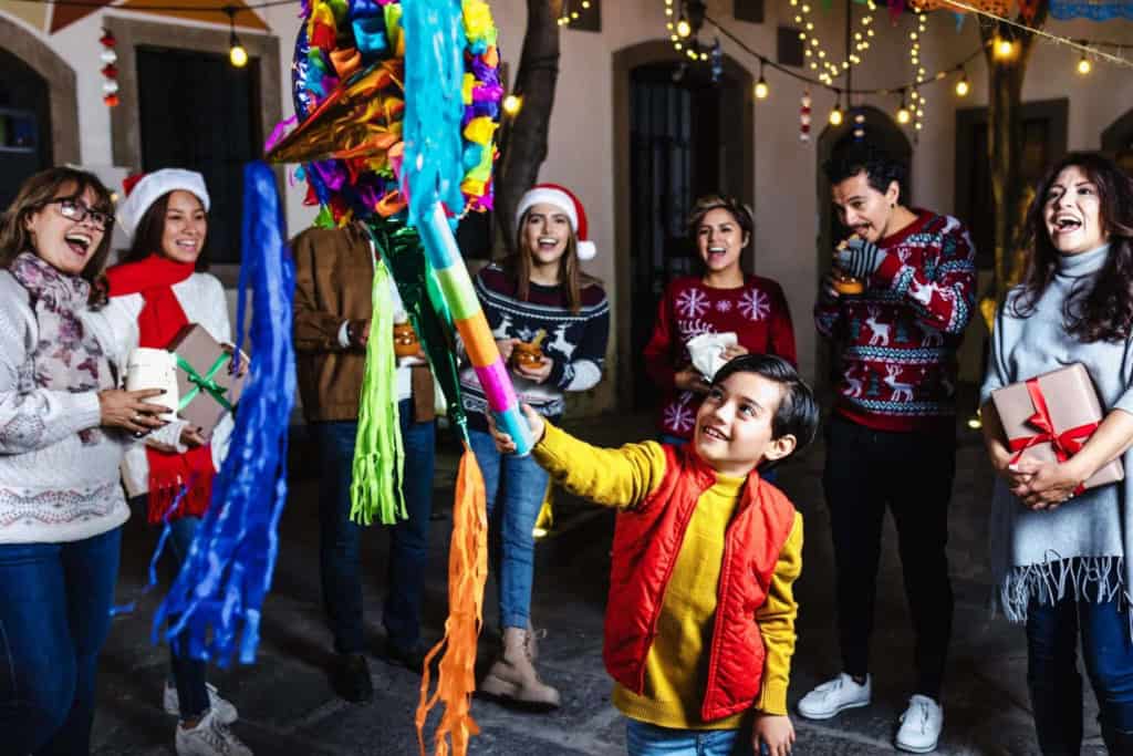 A joyful group of children and adults celebrating Christmas outdoors, exchanging gifts, and enjoying festive decorations and colorful lights during a holiday gathering.