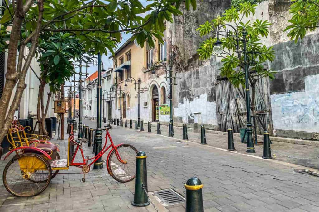 An empty street in Indonesia featuring traditional buildings, lush greenery, and a colorful bicycle rickshaw parked along the sidewalk. The scene captures the local atmosphere and vibrant culture.