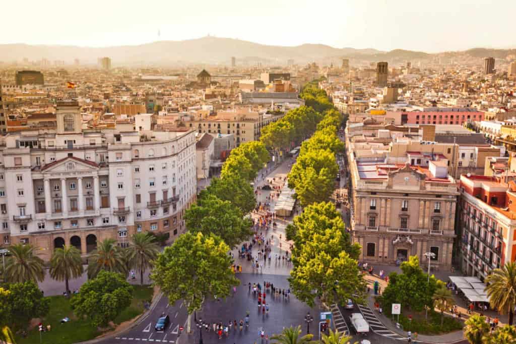 Aerial view of Barcelona showcasing a prominent tree-lined street with bustling activity, historic architecture, and scenic cityscape in the background.