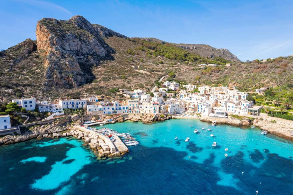 A picturesque coastal village in Italy with white buildings, a small harbor, and boats on clear blue waters, set against a backdrop of rugged hills and a bright, sunny sky.