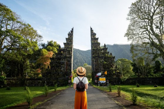 A traveler stands facing a historic temple gate in Indonesia, surrounded by vibrant nature and scenic mountain views, illustrating the cultural richness and natural beauty of the country.