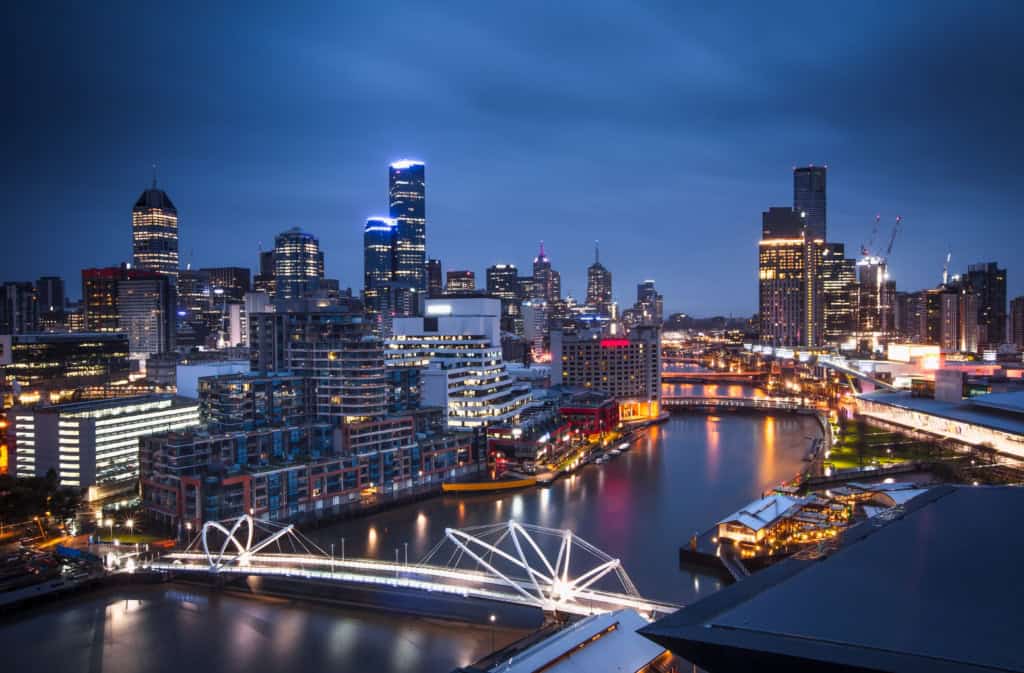 An aerial view of Melbourne's vibrant cityscape during evening hours, showcasing modern skyscrapers, the Yarra River, and city lights reflecting on the water.
