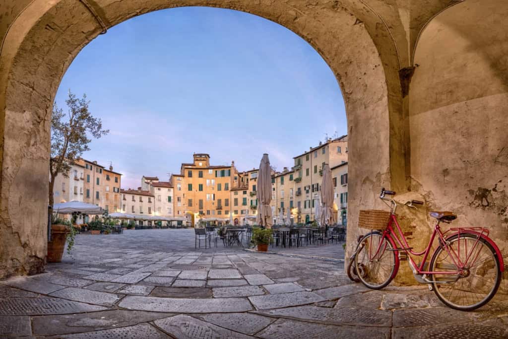 A picturesque view of a historic Italian village square framed by an archway, featuring colorful buildings, outdoor cafes, and a vintage pink bicycle resting against the wall.