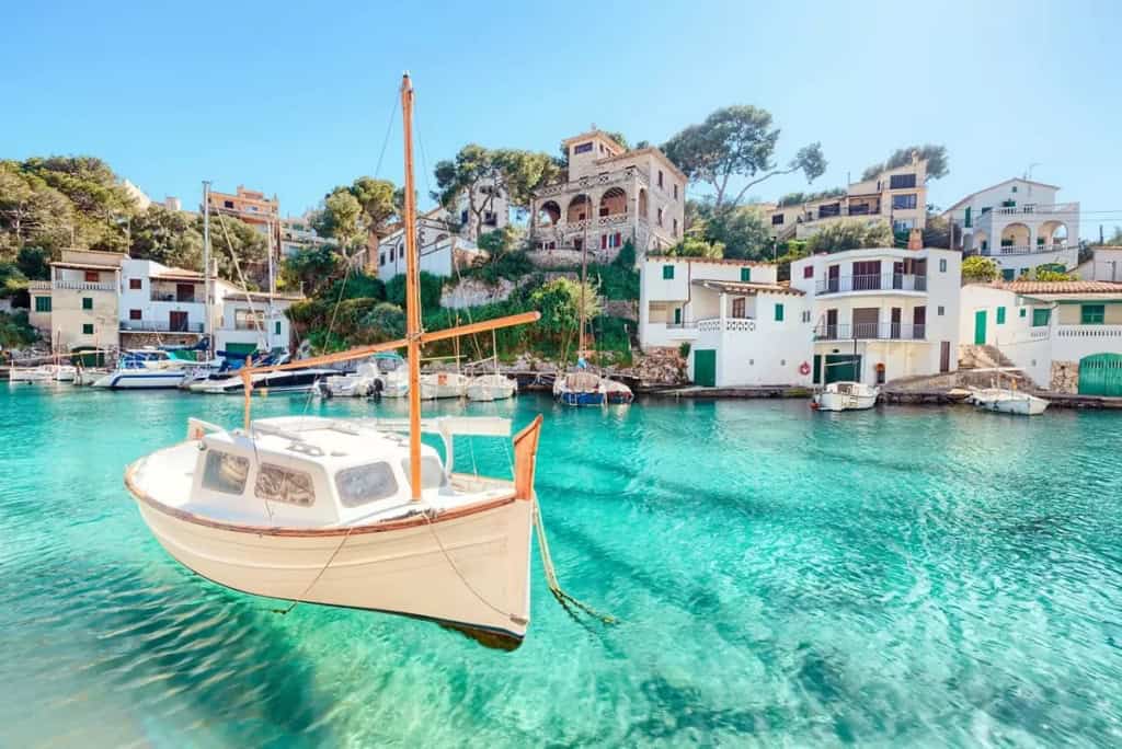 An idyllic scene of a small boat anchored in clear turquoise waters near a picturesque seaside village in Spain. The area features colorful houses and lush greenery, perfect for expatriates seeking a.
