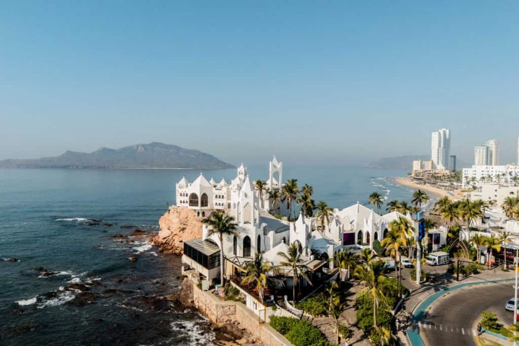 An aerial view of a coastal city in Mexico, showcasing a white church on the rocky shoreline, palm trees, and modern high-rise buildings in the background, highlighting popular retirement destinations.