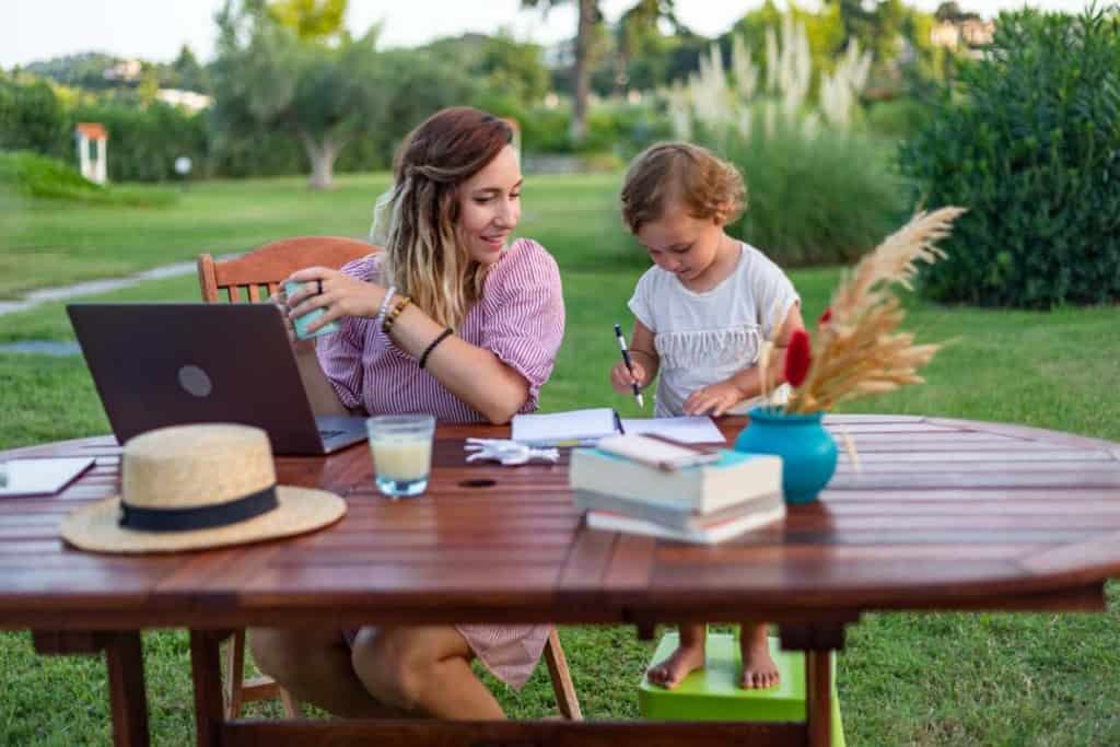 A woman and a young girl enjoying outdoor activities at a table, with a laptop, books, and a glass of water, surrounded by lush greenery, representing expatriate life and family moments in Greece.