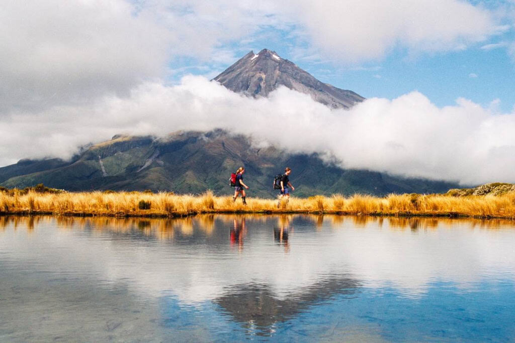 Mount Taranaki with hikers walking along a lake in New Zealand, showcasing the country's stunning natural landscapes and outdoor adventure opportunities.