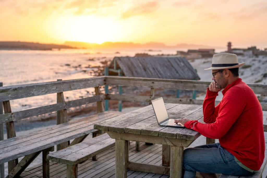 A man working on a laptop outdoors at sunset, with a scenic coastal view and wooden deck, representing international relocation and digital nomad lifestyle.
