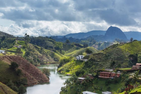A panoramic view of lush green hills, a winding river, and scattered houses under a partly cloudy sky in Colombia.