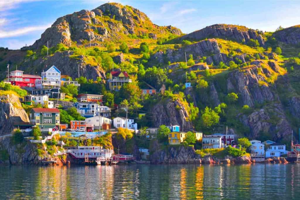 Scenic view of colorful houses on a hillside overlooking a calm water body in a Canadian coastal town, surrounded by rocky cliffs and lush greenery.