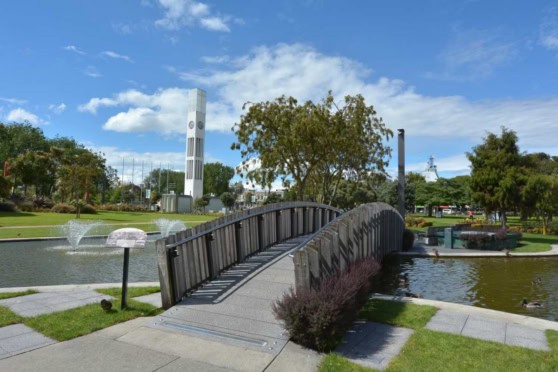 A charming park scene featuring a small, arched wooden bridge crossing a tranquil pond, surrounded by lush greenery and trees under a bright blue sky.