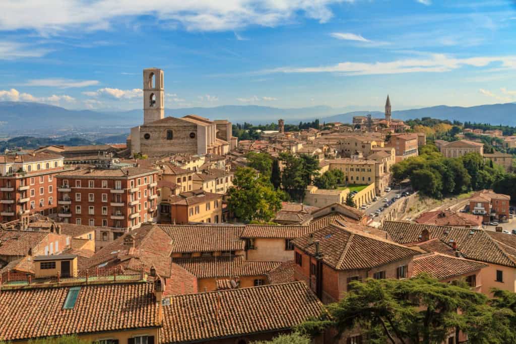An aerial view of a picturesque Italian city featuring historic buildings, terracotta rooftops, and a prominent church tower against a backdrop of rolling hills and a bright blue sky.