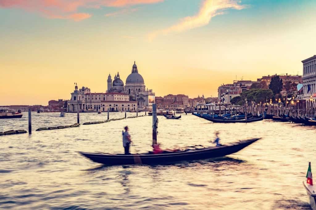 A gondolier navigates a gondola through Venice's canals during sunset, with historic buildings and the Basilica di Santa Maria della Salute in the background.