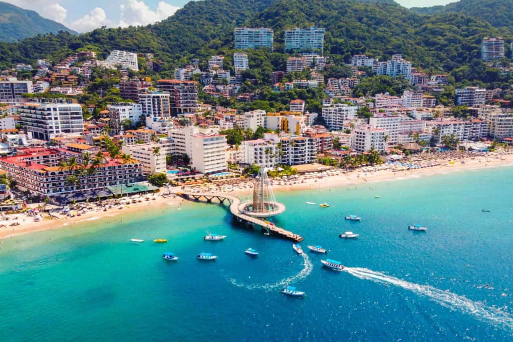 Scenic view of a Mexican seaside town with colorful buildings, a sandy beach, and boats anchored in the calm, blue ocean.