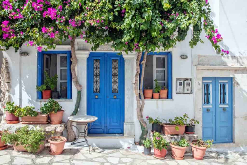 Beautiful Greek home featuring vibrant blue doors and window frames, surrounded by lush potted plants and colorful flowers, embodying the picturesque charm of Greece.