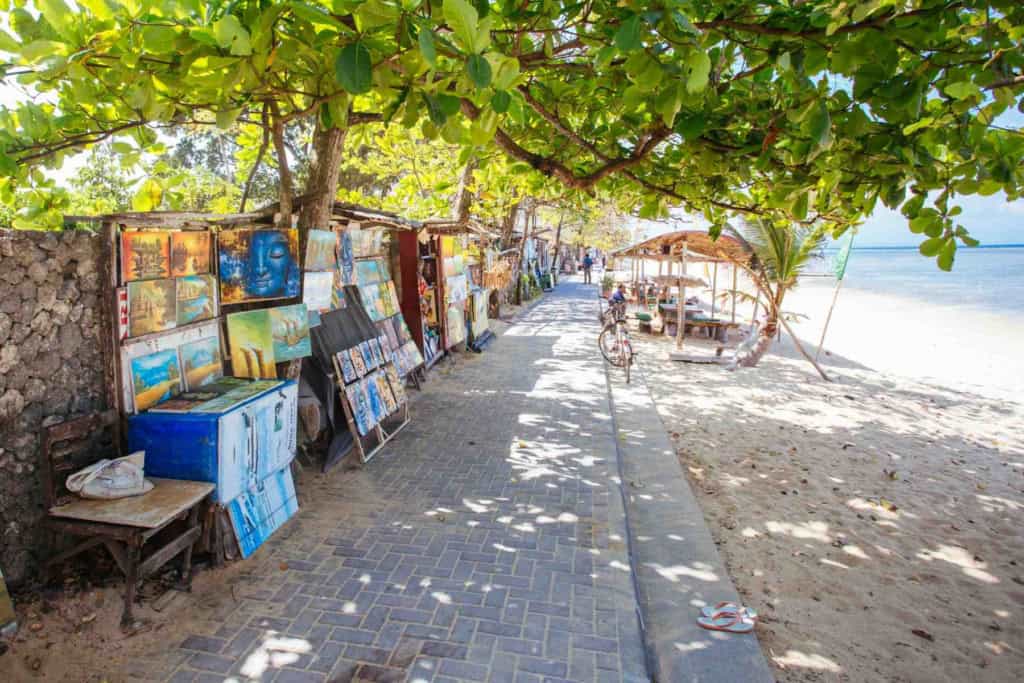 Colorful artworks displayed along a shaded pathway by the beach, showcasing local artists' creations in a lively, scenic setting.
