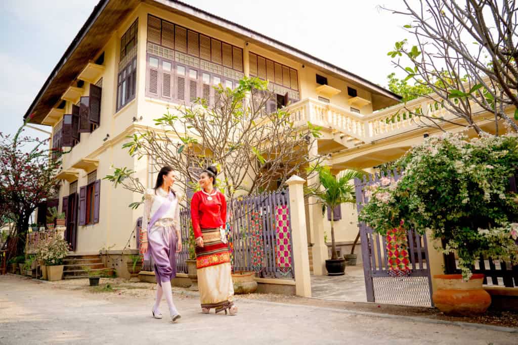Two women in traditional Thai attire chatting outside a charming Thai house, illustrating cultural integration and lifestyle in Thailand.