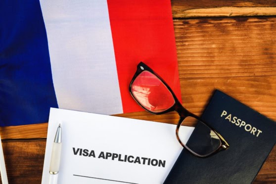 An image showing a visa application form, passport, glasses, and a pen on a wooden surface with a French flag in the background, representing international travel and visa procedures.