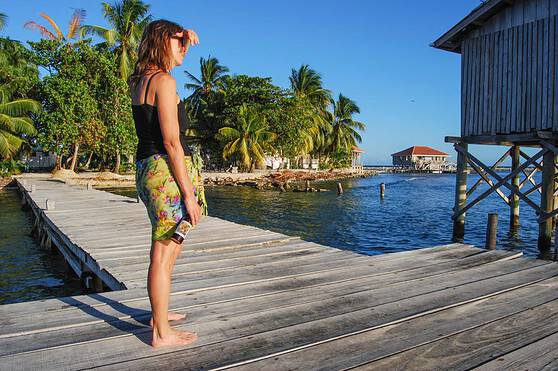 A woman standing on a wooden dock overlooking the clear blue waters of Belize, surrounded by lush palm trees and tropical scenery, illustrating a peaceful Caribbean lifestyle.