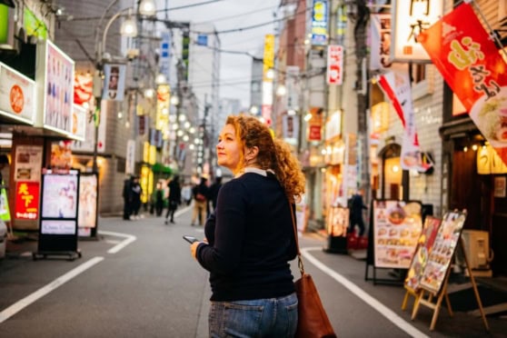 An Asian woman with curly hair exploring a lively street in Japan, surrounded by colorful signs, shops, and restaurants, capturing the energetic atmosphere of urban Japan.