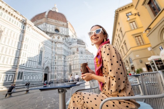 An elegant woman with sunglasses and a polka-dot dress sitting at an outdoor café in Florence, Italy, with the Florence Cathedral (Duomo) in the background, capturing a moment of leisure and travel.