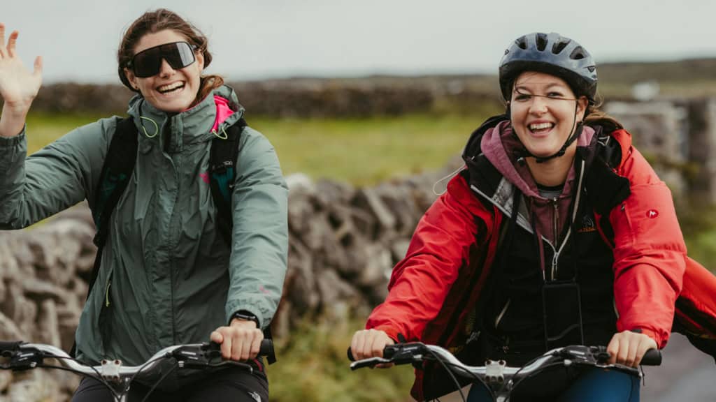 Two women smiling and riding bicycles outdoors on a scenic trail, dressed in outdoor gear with helmets, enjoying a sunny day and the freedom of cycling in nature.