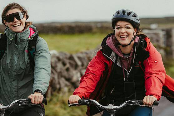Two women smiling and riding bicycles outdoors on a scenic trail, dressed in outdoor gear with helmets, enjoying a sunny day and the freedom of cycling in nature.
