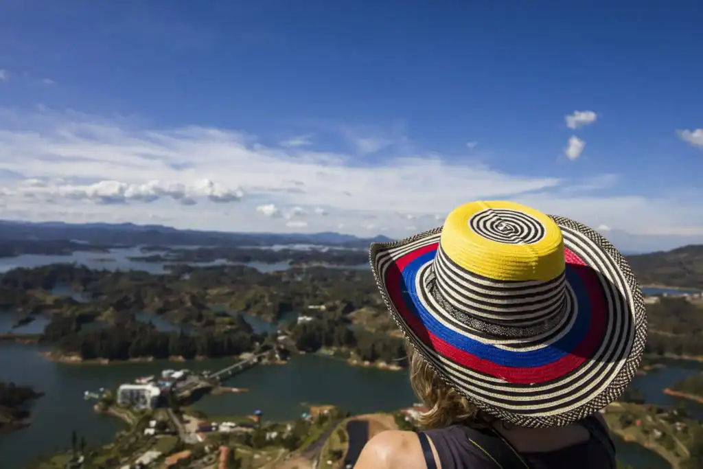 An aerial view of Colombia's lush landscape featuring lakes, forests, and mountains, with a woman wearing a colorful striped hat enjoying the breathtaking scenery.