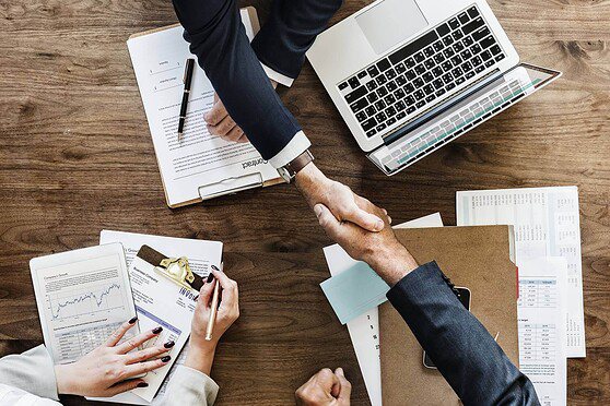 Business professionals shaking hands over a wooden table with documents, laptop, and charts, representing international insurance services and global business partnerships.