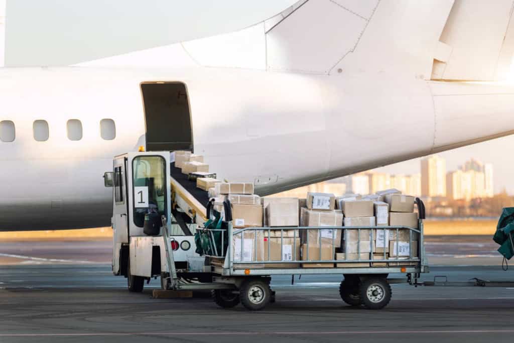 An airplane on the tarmac with a baggage cart filled with luggage being loaded into the aircraft's cargo hold. The scene is set during daylight with a city skyline in the background.