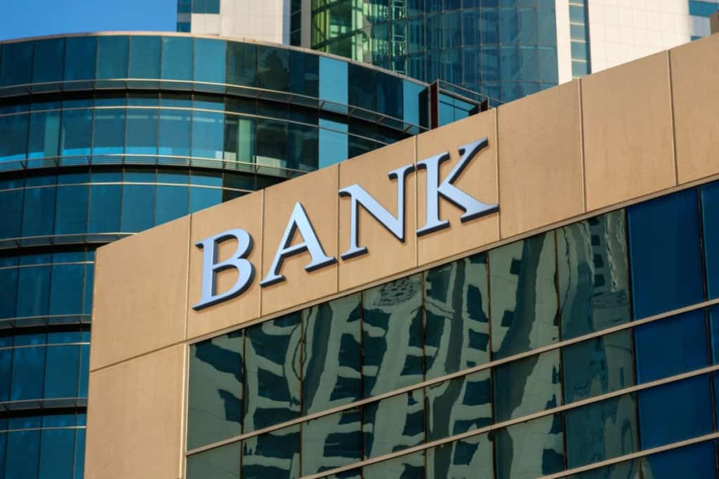 Modern bank building with glass facade and prominent "BANK" sign, reflecting urban financial district surroundings.