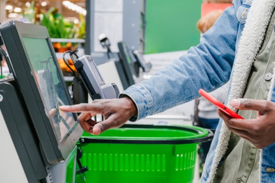 Customer using self-checkout kiosk at a grocery store, scanning items with a smartphone in hand.