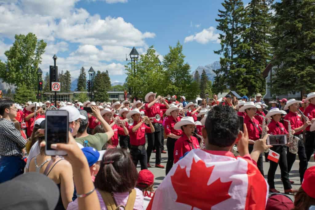 Diverse crowd participating in a lively outdoor parade with Canadian flags, surrounded by trees and mountains under a bright blue sky.