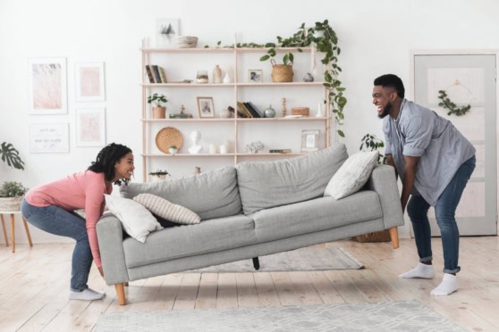 Two people moving a gray sofa in a bright, modern living room with white walls, wooden flooring, and decorative shelves with plants and books.
