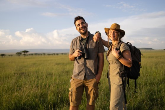 Happy couple enjoying outdoor adventure in a vast grassy field, equipped with backpacks and binoculars, under a bright sky with scattered clouds, showcasing travel and exploration.