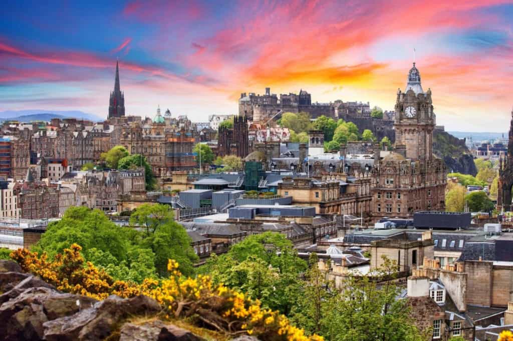 Panoramic view of Edinburgh's historic cityscape during sunset, showcasing iconic landmarks like Edinburgh Castle and the Scott Monument with vibrant sky colors.