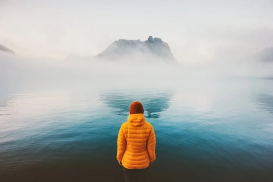 A person in a yellow jacket standing alone by a calm lake with misty mountains in the background, symbolizing feelings of isolation and contemplation for expats dealing with depression.