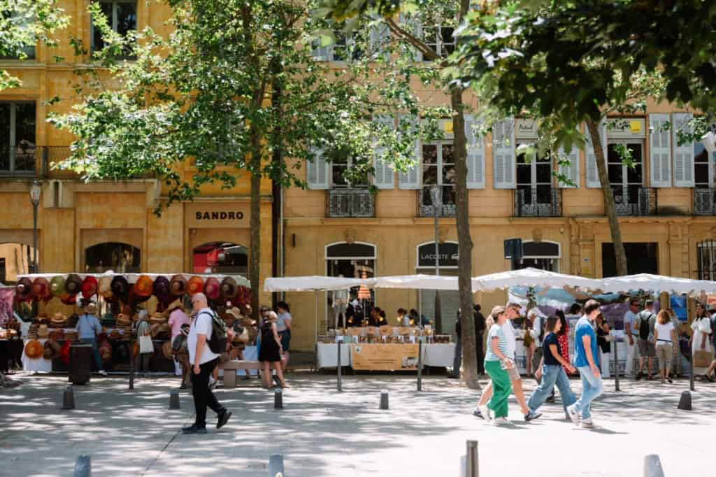 People walking past outdoor market stalls in a French city with historic buildings and trees.
