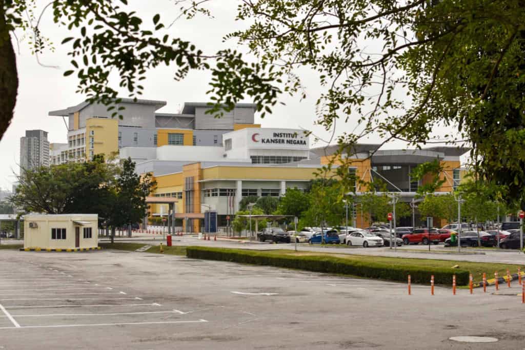 An exterior view of a modern healthcare institute in Malaysia, featuring a large building with medical signage, surrounded by trees and parking spaces, ideal for retirees seeking quality medical servi.