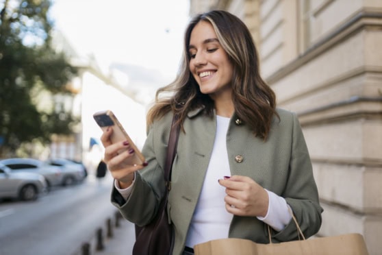 A woman with long brown hair, wearing a green jacket and white top, smiling while looking at her smartphone on a city street.