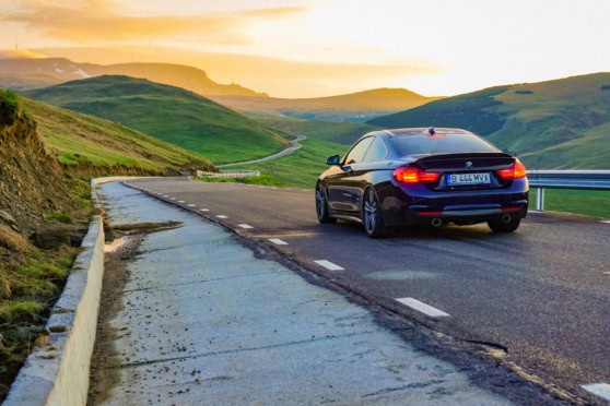 A sleek black luxury car driving along a winding mountain road during sunset, surrounded by lush green hills and a beautiful landscape.