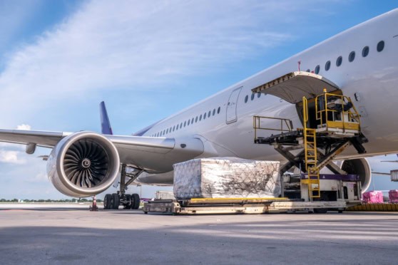 Cargo being loaded onto a commercial airplane at an airport, with a cargo lift and pallet, under a clear blue sky.