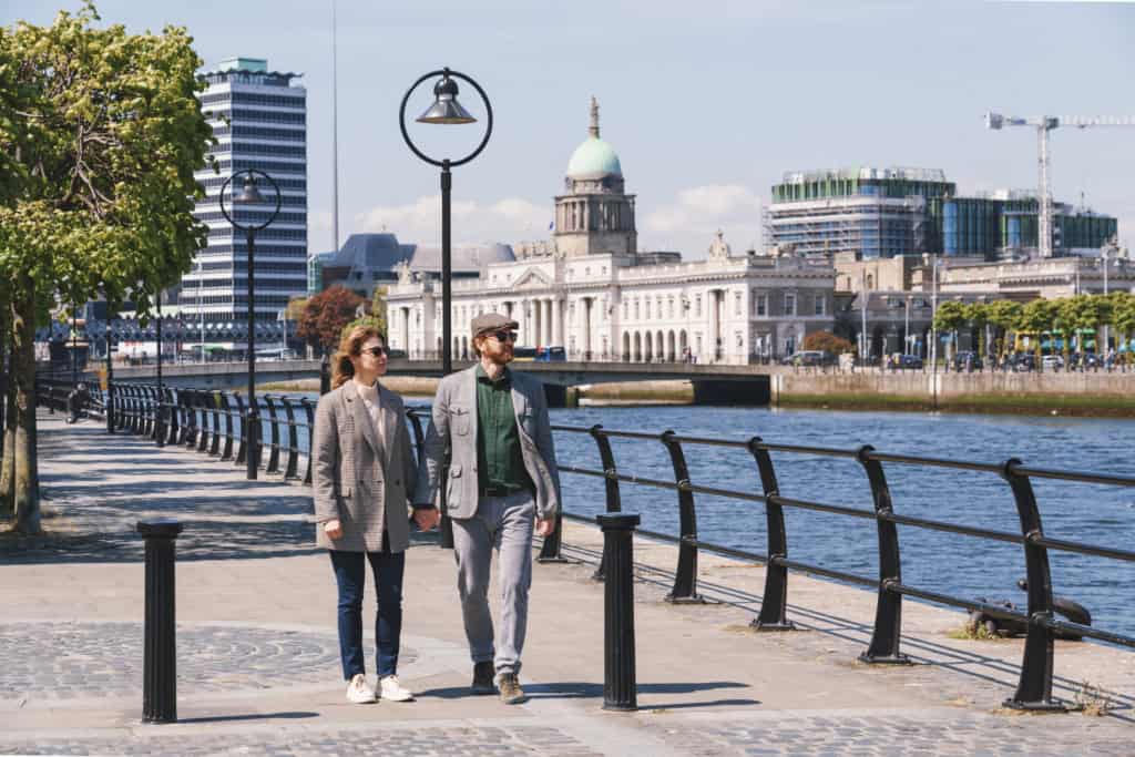 A couple walking along the Thames River in London, with historic and modern buildings in the background, under a clear blue sky.