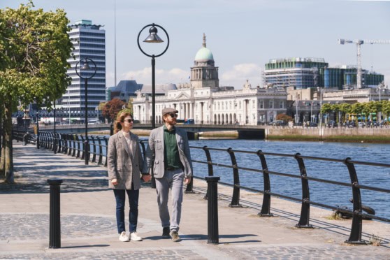 A couple walking along the Thames River in London, with historic and modern buildings in the background, under a clear blue sky.
