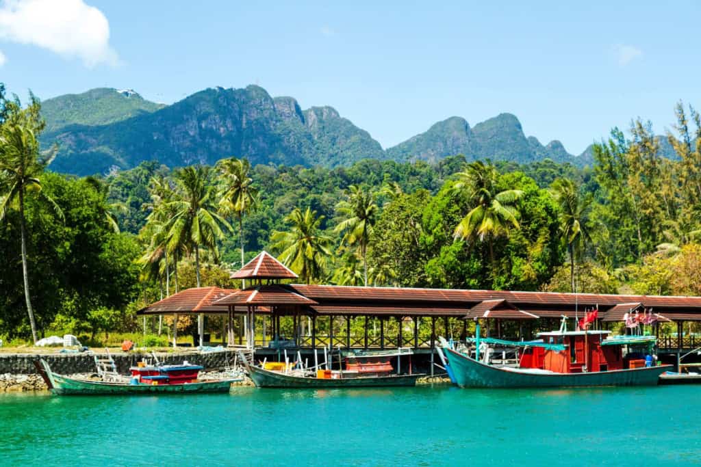 A scenic view of boats docked at a waterfront with lush green mountains in the background, ideal for retirees seeking a tropical paradise in Malaysia.