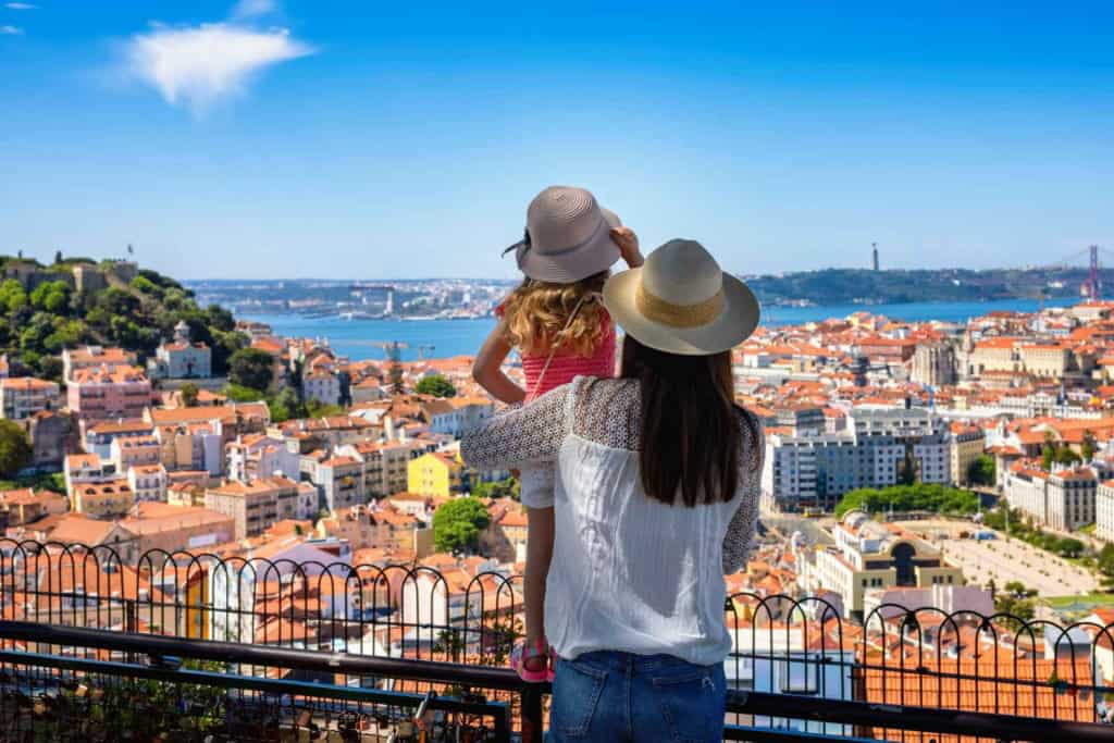 A woman and a girl enjoying a panoramic view of a vibrant city with historic buildings, a river, and a bridge in the background, on a sunny day.