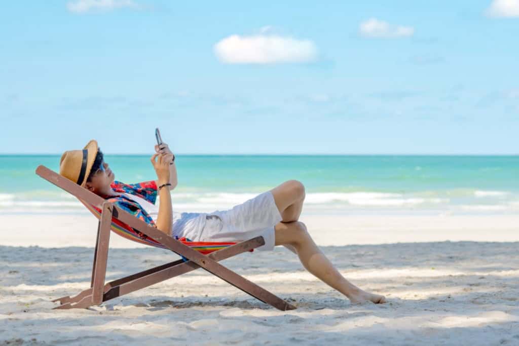 Relaxing man on a beach chair using a smartphone and researching the best international credit cards, with the ocean and blue sky in the background.