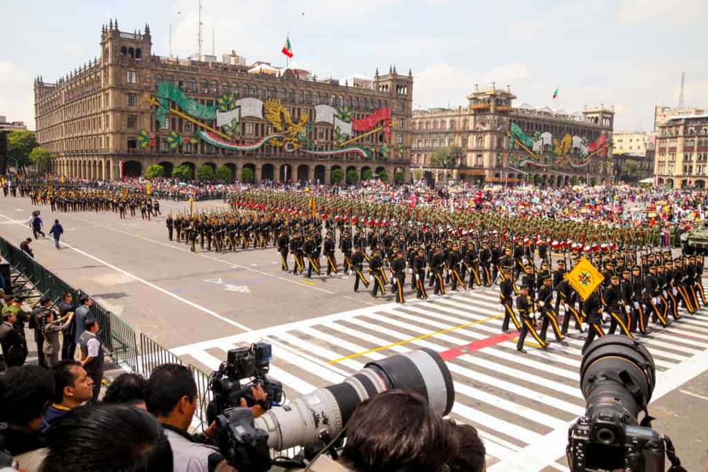A large parade with military personnel marching in formation across a city square, with a historic building decorated with colorful banners in the background.